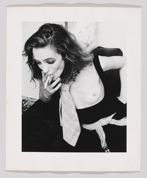 A woman with one breast exposed leans forward and smokes a cigarette while wearing a tie.