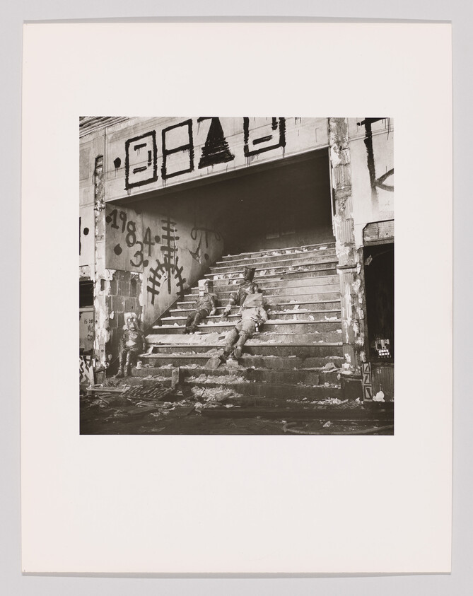 Two people sit slumped on debris-strewn steps inside a graffiti-covered, damaged building.