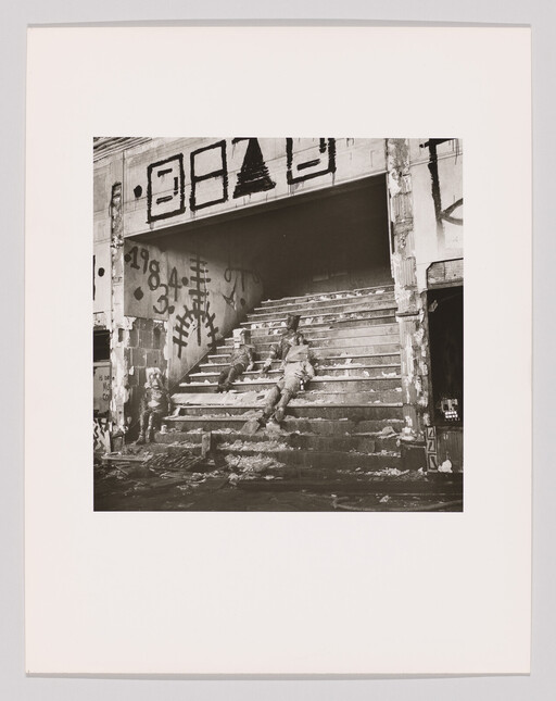 Two abandoned, injured-looking figures slumped on debris-covered stairs inside a graffiti-covered, ruined building.