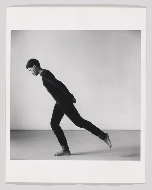 A barefoot dancer in a black outfit leans forward with hands clasped and one leg extended behind.