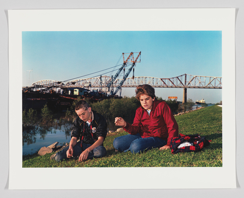 Two young individuals are sitting on a grassy riverbank with a large metal bridge in the background. The person on the left is focused on the ground, possibly examining something, while the person on the right is looking intently at an object in their hand. The setting appears to be in the late afternoon with clear skies.