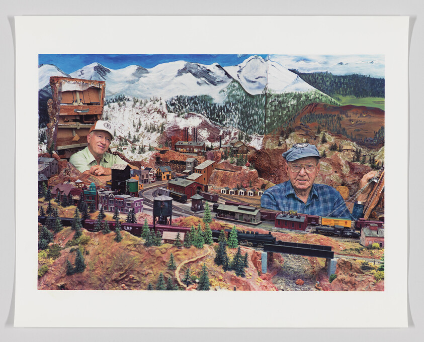 A detailed model train set with miniature buildings, trees, and trains is depicted, with snowy mountains in the background. Two cut-out photos of smiling men, one younger and one older, are superimposed onto the scene, giving the playful impression that they are giants within the miniature landscape.