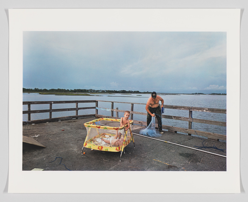 A photograph depicting a shirtless man standing on a wooden pier by the water, holding a fishing net, with a young child seated in a colorful playpen nearby. The sky is overcast and the pier overlooks a calm body of water with land visible in the distance.