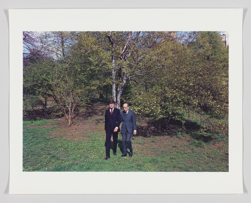 Two men in suits and hats standing on a grassy field with trees in the background on a sunny day. The image is framed with a white border.