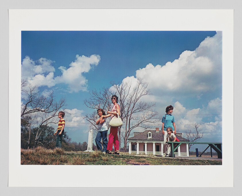 A group of people, including children and adults, walking in a line across a grassy field with a clear blue sky and fluffy clouds overhead. In the background, there is a small house with a porch and a bridge to the right. The image has a vintage look, suggesting it may have been taken in the mid-20th century.