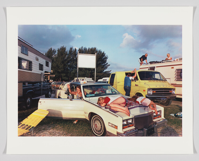 A vibrant scene at a drive-in theater with various people and vehicles. A woman lounges on the hood of a white car, two children sit inside the car looking out, a man in a blue shirt leans on a yellow van, and two people sit on top of an RV enjoying the view. The atmosphere is casual and relaxed with a clear sky above.