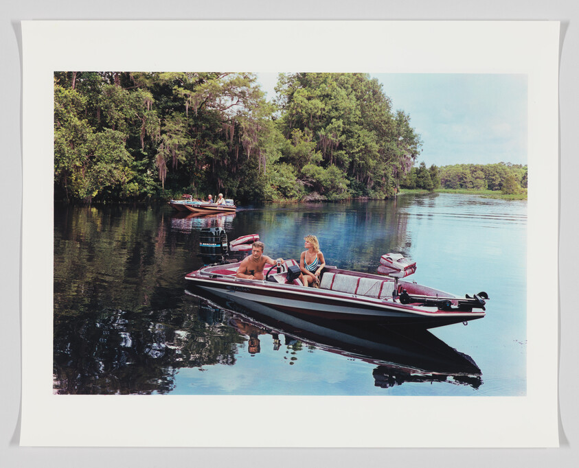 A serene river scene with two speedboats carrying people, one in the foreground with a man and a woman, and another in the background with several passengers. The river is surrounded by lush greenery and trees with Spanish moss, reflecting the clear blue sky above.