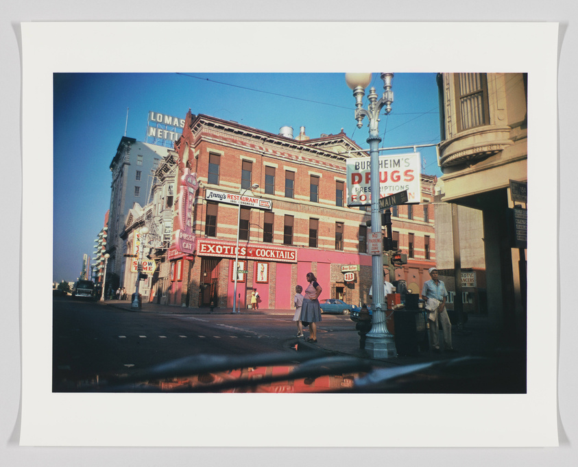 A vintage street scene with old buildings adorned with various signs, including "Exotics & Cocktails" and "Burheim's Drugs." Pedestrians are walking on the sidewalk, and the image has a clear blue sky above. The photo has a white border, suggesting it may be a print or developed photograph.