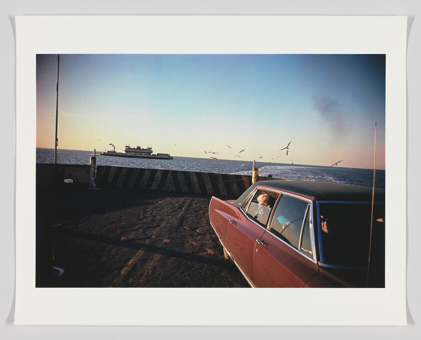 A vintage red car parked on a pier with a couple inside looking out to sea where a ferry is visible in the distance. Seagulls are flying in the sky above the water, and the scene is set against a clear sky with a hint of sunset. The image is framed with a white border.