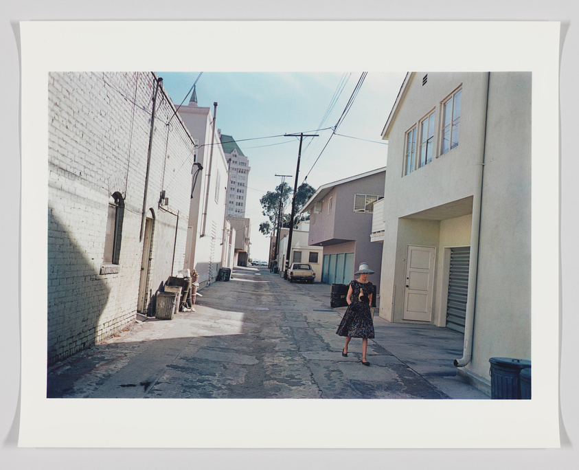 A photograph of a woman walking down an urban alleyway with buildings on either side and a clear blue sky above. The alley is lined with various objects like trash bins and a parked van. The image has a white border, suggesting it is a print or a photograph of a photograph.