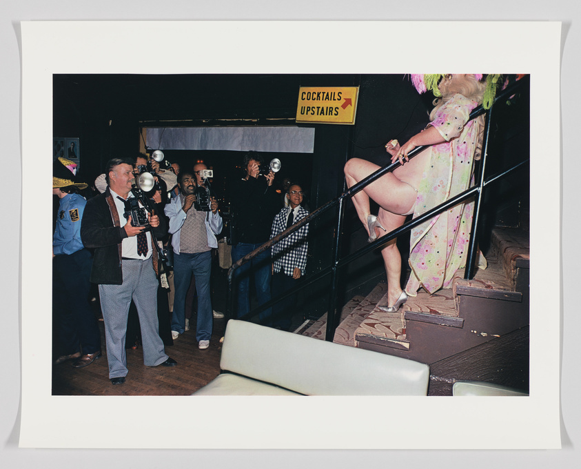 A group of photographers with cameras and flash equipment are taking photos of a person in a flamboyant costume ascending a staircase next to a sign that reads "COCKTAILS UPSTAIRS." The person is wearing a dress with a floral pattern, a green wig, and high heels. The scene appears to be in a club or event space, and there is a mix of amusement and surprise on the faces of the onlookers.