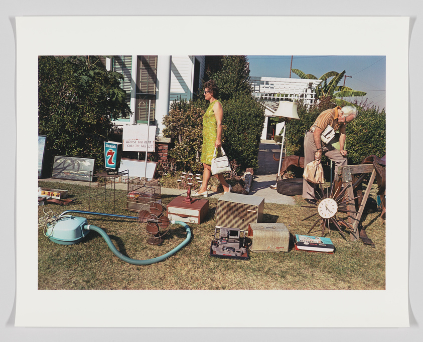 A woman and a man browse items at a yard sale in front of a house on a sunny day. The woman, dressed in a yellow dress, carries a white handbag and walks past various objects scattered on the lawn, including a vacuum cleaner, cages, and a clock. The man, wearing a striped shirt and glasses, examines an item closely. A sign reading "House for Rent" is visible in the background.