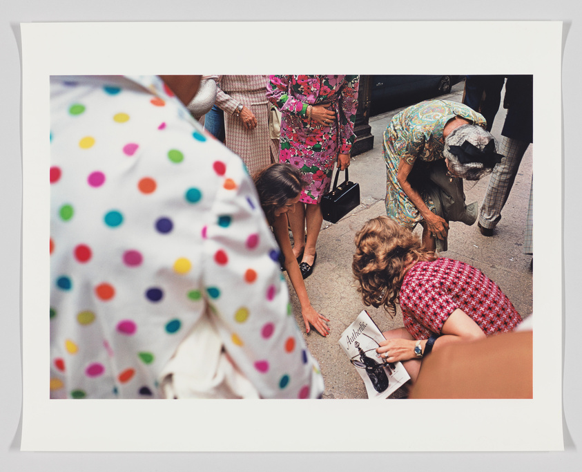 A candid street scene with several people, predominantly women, gathered around a dropped item. A woman in a red dress with a houndstooth pattern is bending over to pick up a book titled "Autobiography," while others in colorful, patterned clothing look on or assist. The foreground is partially obscured by a person wearing a white garment with multicolored polka dots.