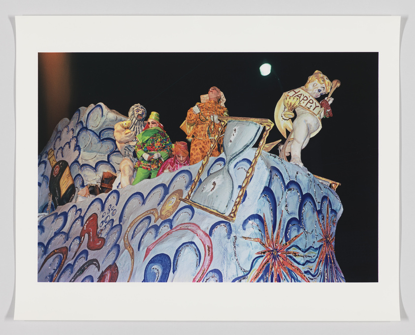 A vibrant and colorful float featuring costumed figures and a large, whimsical wave design, with a figure holding a sign that reads "HAPPY" at a nighttime parade or carnival event.