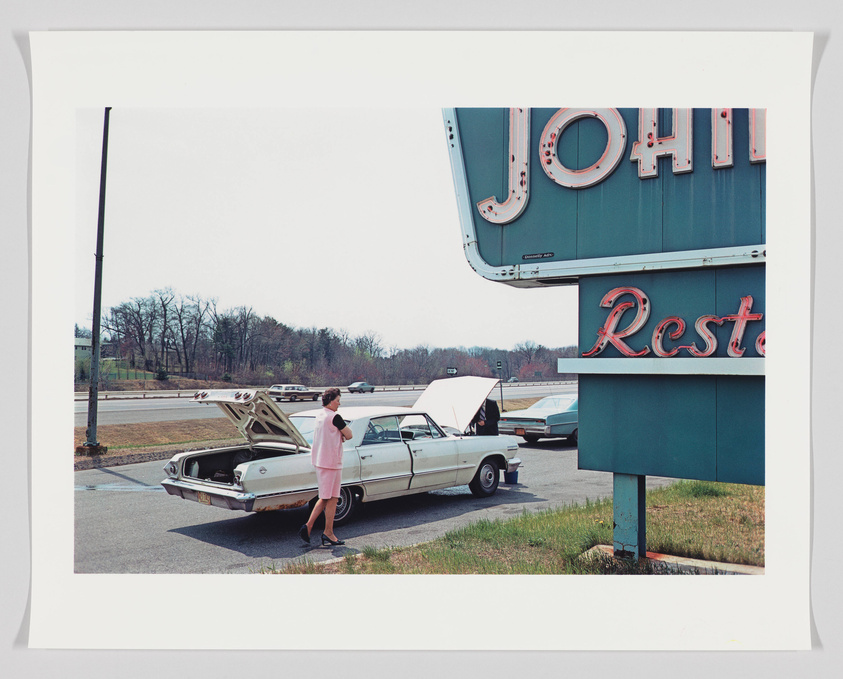 A vintage scene with a person standing next to an open trunk of a classic car in front of a retro diner sign that reads "JOHN'S Restaurant." The setting appears to be a parking lot with another car and a highway in the background.