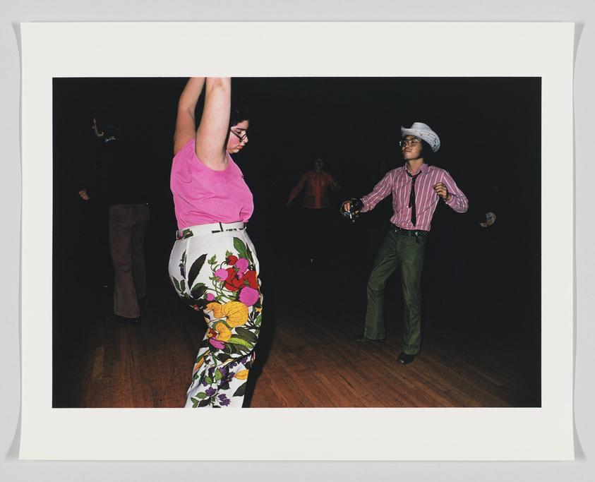 A photograph capturing two people dancing on a wooden floor. The person on the left is wearing a bright pink top and floral pants, with one arm raised. The person on the right, wearing a striped shirt, bell-bottom pants, and a white cowboy hat, is dancing with a tambourine in hand. Other dancers are partially visible in the background, all set against a dark backdrop.