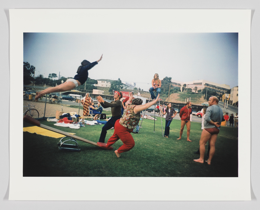 A vintage photograph capturing a lively outdoor scene with a person in mid-air performing a backflip on a trampoline, surrounded by onlookers in various casual and athletic attire, with a mix of concern and amusement on their faces. In the background, there's a grassy field, a fence, and buildings under a cloudy sky.