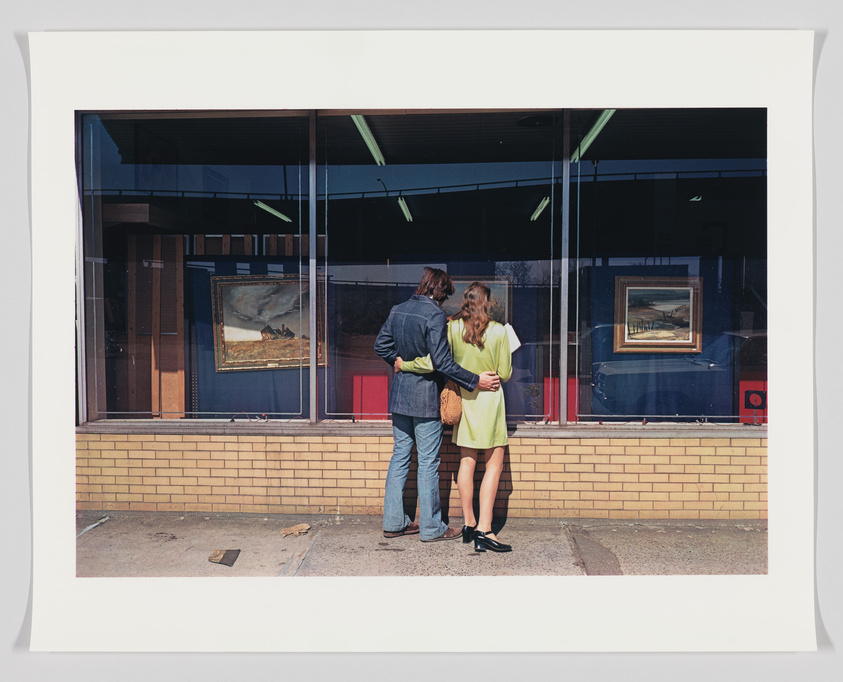 A couple stands closely together in front of a large window displaying paintings, with the man's arm around the woman. The window reflects the blue sky and some interior elements, while the exterior wall features a brick base. The image is framed with a white border.