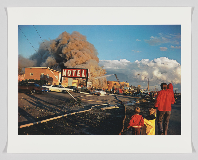 A large plume of smoke rises from behind a two-story building with a "MOTEL" sign in the foreground. Onlookers, including a family with two children, watch the scene from across the street under a clear sky with puffy clouds. Firefighting efforts are visible with a fire truck and a ladder extended towards the smoke. The scene is bathed in the warm glow of the sun, casting long shadows on the pavement.