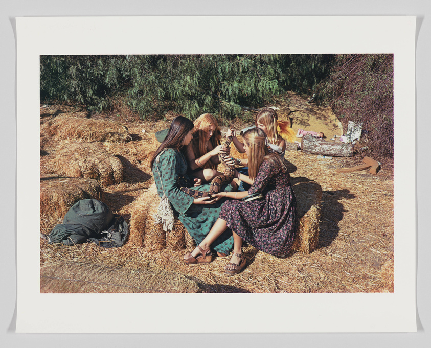 Three women sitting on hay bales engaging in a craft activity outdoors, with a backpack on the ground next to them and a child lying on a blanket in the background.