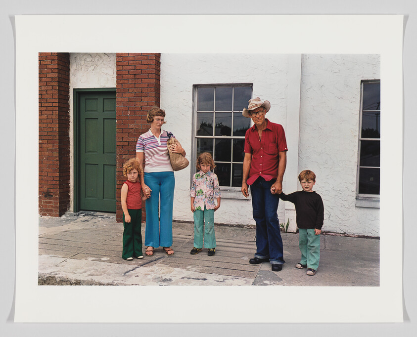 A vintage photograph of a family of five standing in front of a white building with a green door and a window. From left to right: a young girl in a red sleeveless top and green pants, a woman holding a purse wearing a striped shirt and blue pants, another young girl in a patterned shirt and green pants, a man in a red shirt and jeans with a cowboy hat, and a young boy in a brown shirt and green pants. They all have serious expressions and are looking directly at the camera.