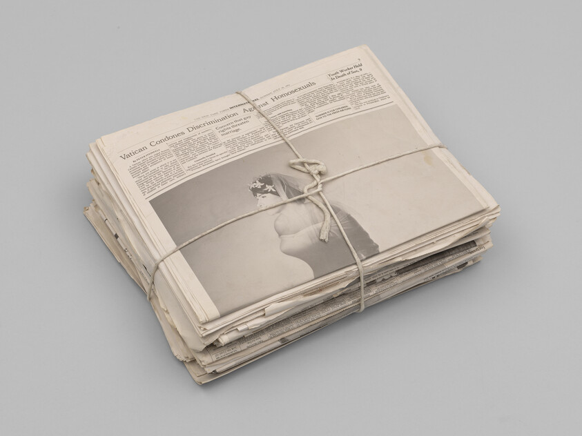 A stack of aged newspapers tied together with string, placed on a solid grey background. The visible top newspaper features a headline about the Vatican and an image of a classical statue.