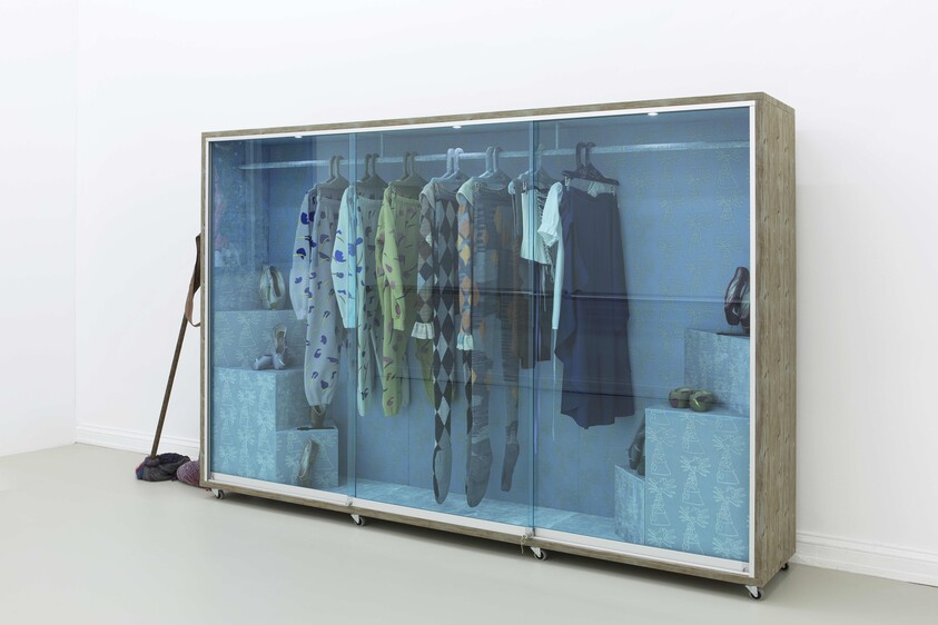 A wooden wardrobe with blue-tinted glass doors displays hanging clothes, shoes, and folded boxes inside.