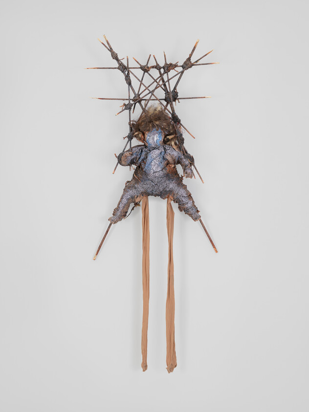 A hanging mixed-media sculpture resembling a seated, spiky figure with long tan fabric legs.