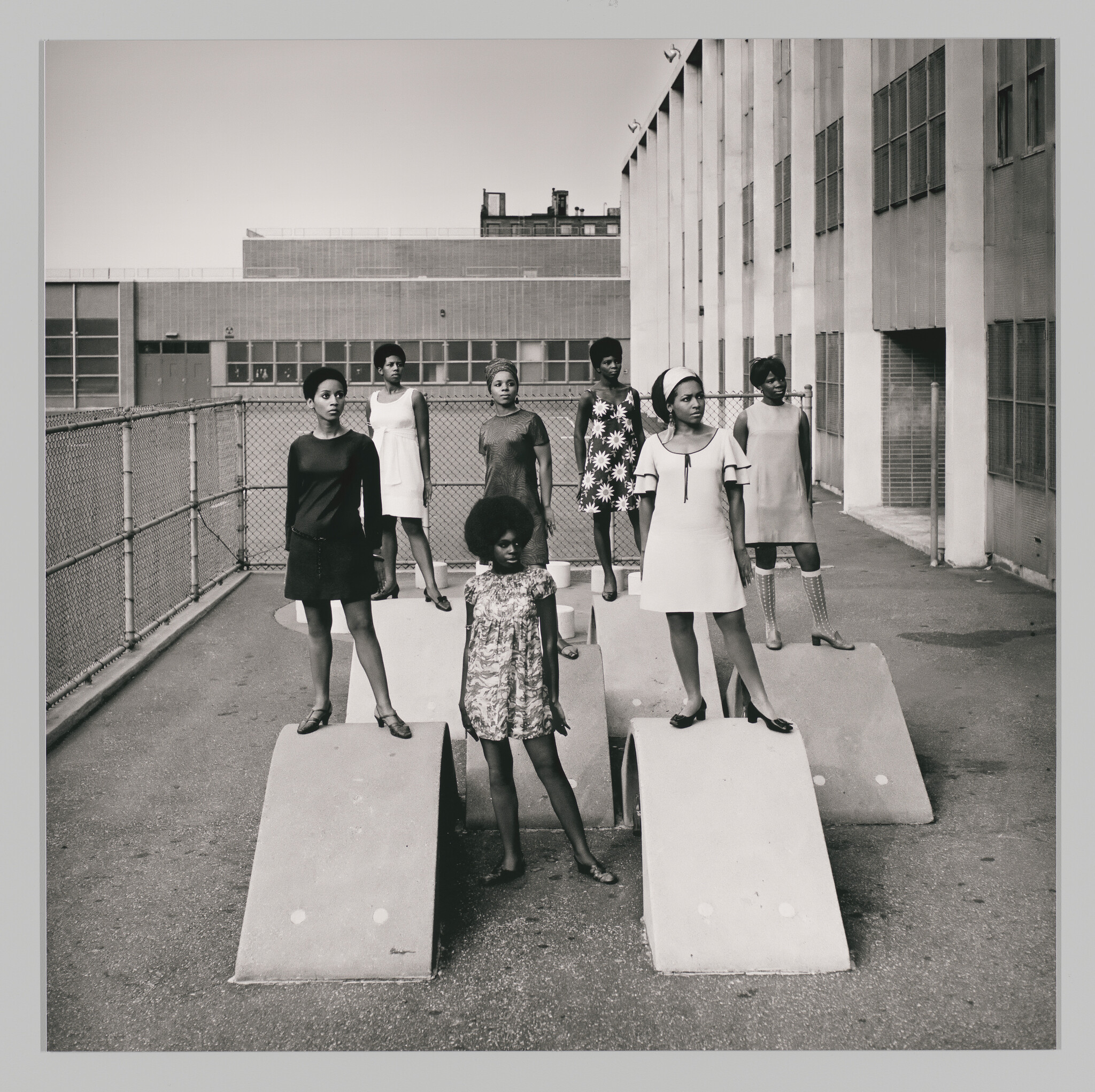 Seven young women stand confidently on concrete arches in a fenced urban schoolyard.