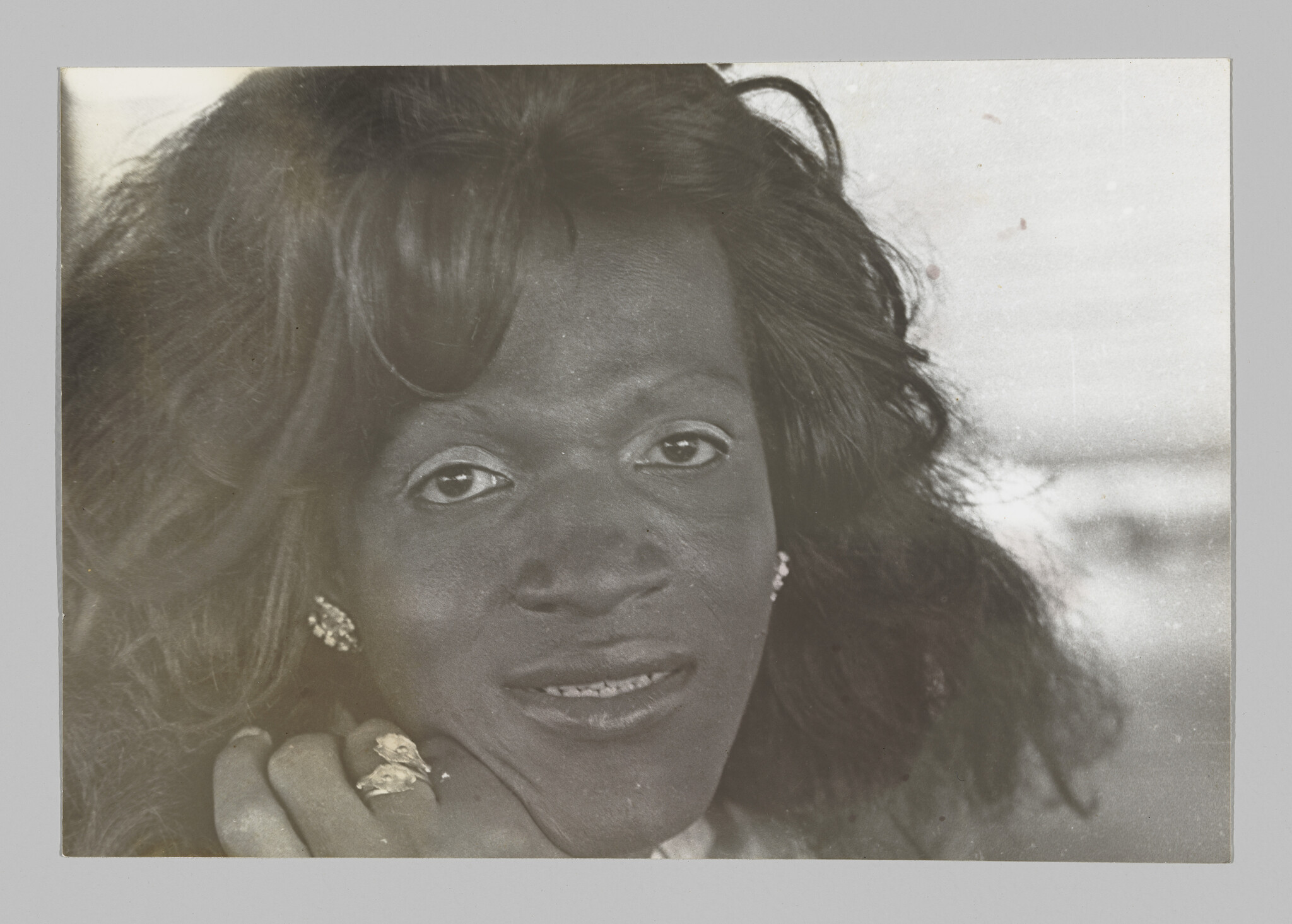 Closely cropped portrait of a Black trans person gazing into the camera, with shoulder-length hair and glittering jewelry