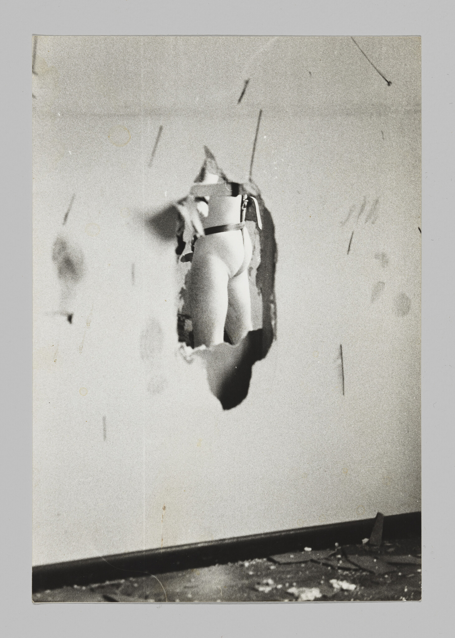 A hole in a dilapidated wall reveals a nude man’s lower back, upper legs, and semi-erect penis from a three-quarter rear view.