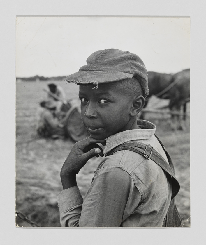 A black and white photograph of a young boy looking over his shoulder at the camera. He wears a worn cap and a button-up shirt with one strap of his overalls hanging loose. In the blurred background, another person is seated on the ground near a bicycle.