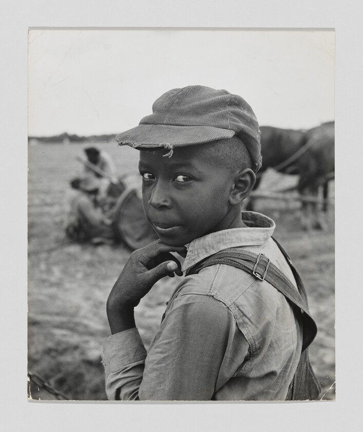 A young boy in a cap and overalls looks over his shoulder in a farm field.