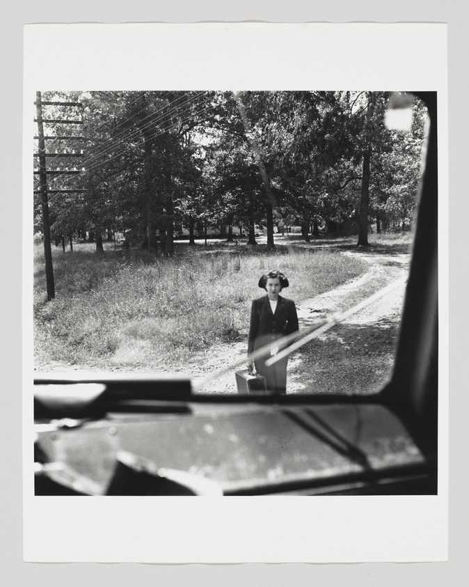 A black and white photograph taken from the inside of a vehicle, showing a woman standing in the center of the frame on a dirt path amidst a grassy area with trees. The woman is dressed in a dark jacket and skirt, with a light-colored blouse, and is holding a suitcase. The car's dashboard and a part of the steering wheel are visible in the foreground, slightly out of focus. Power lines and poles are visible in the background to the left.