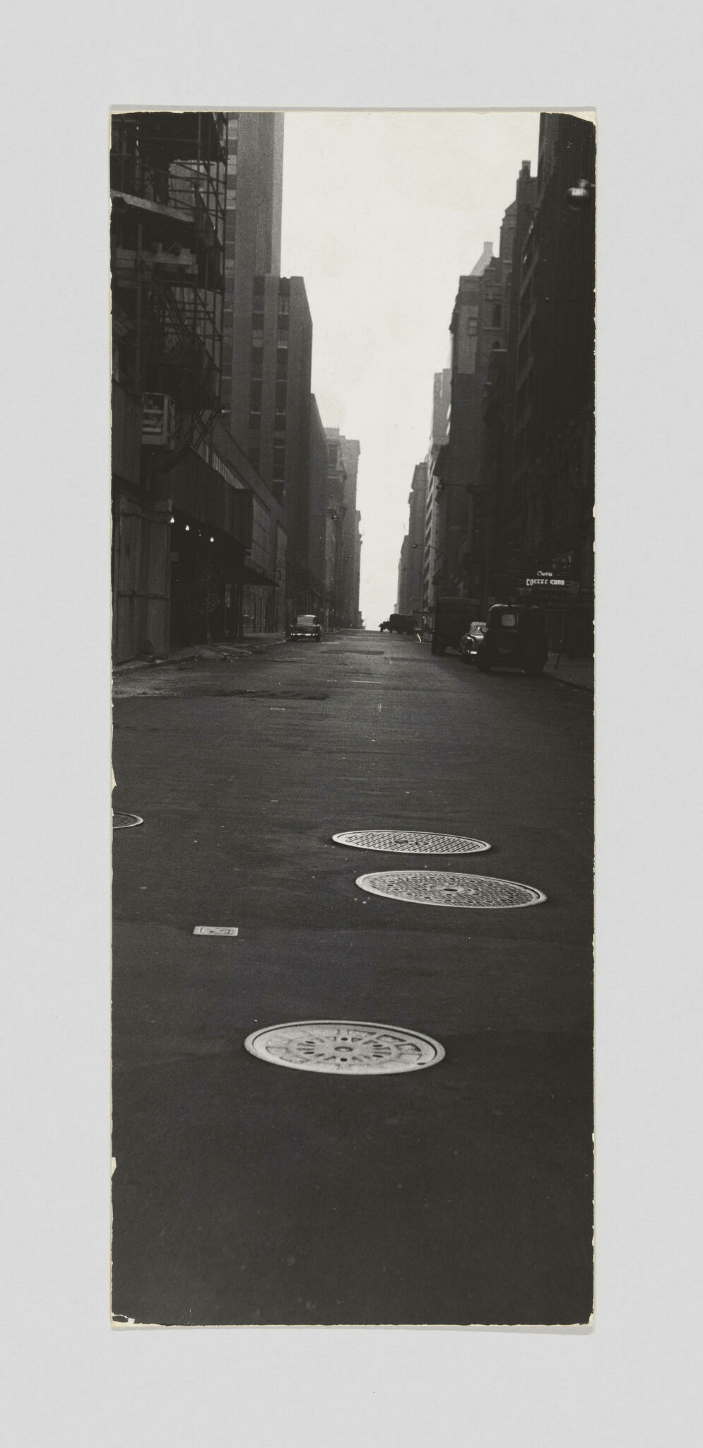 A black and white photograph depicting a deserted urban street flanked by tall buildings, with manhole covers in the foreground and a faintly visible skyline in the distance. The image has a vintage feel, with the edges of the photo showing slight wear.