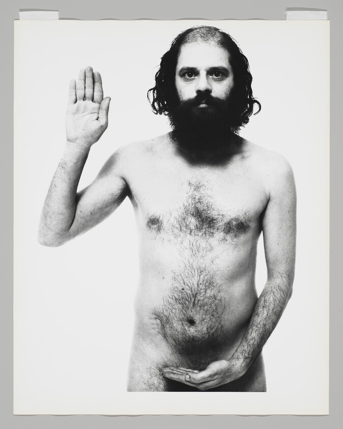 Bearded naked man standing with right hand raised and left hand cupped over his groin.