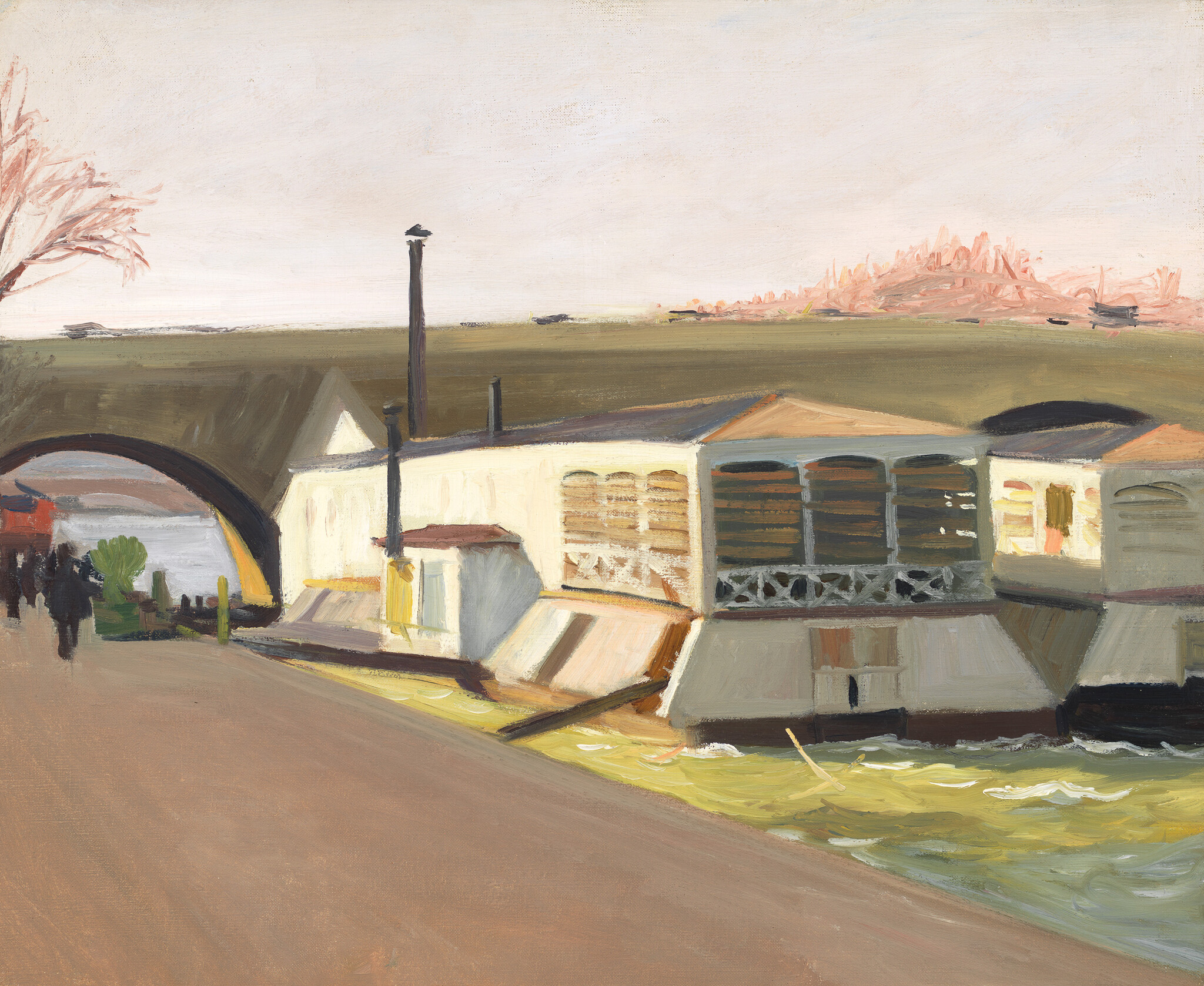A row of riverside boathouses and a walking path under an arch bridge with a few pedestrians.