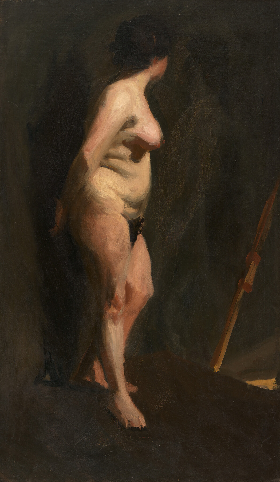A nude woman stands turned slightly away from the viewer in dim studio light.