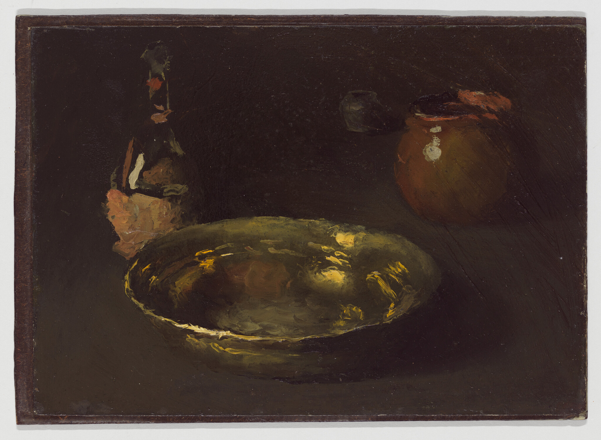 A shiny brass bowl sits on a dark table with a bottle and a clay jar nearby.