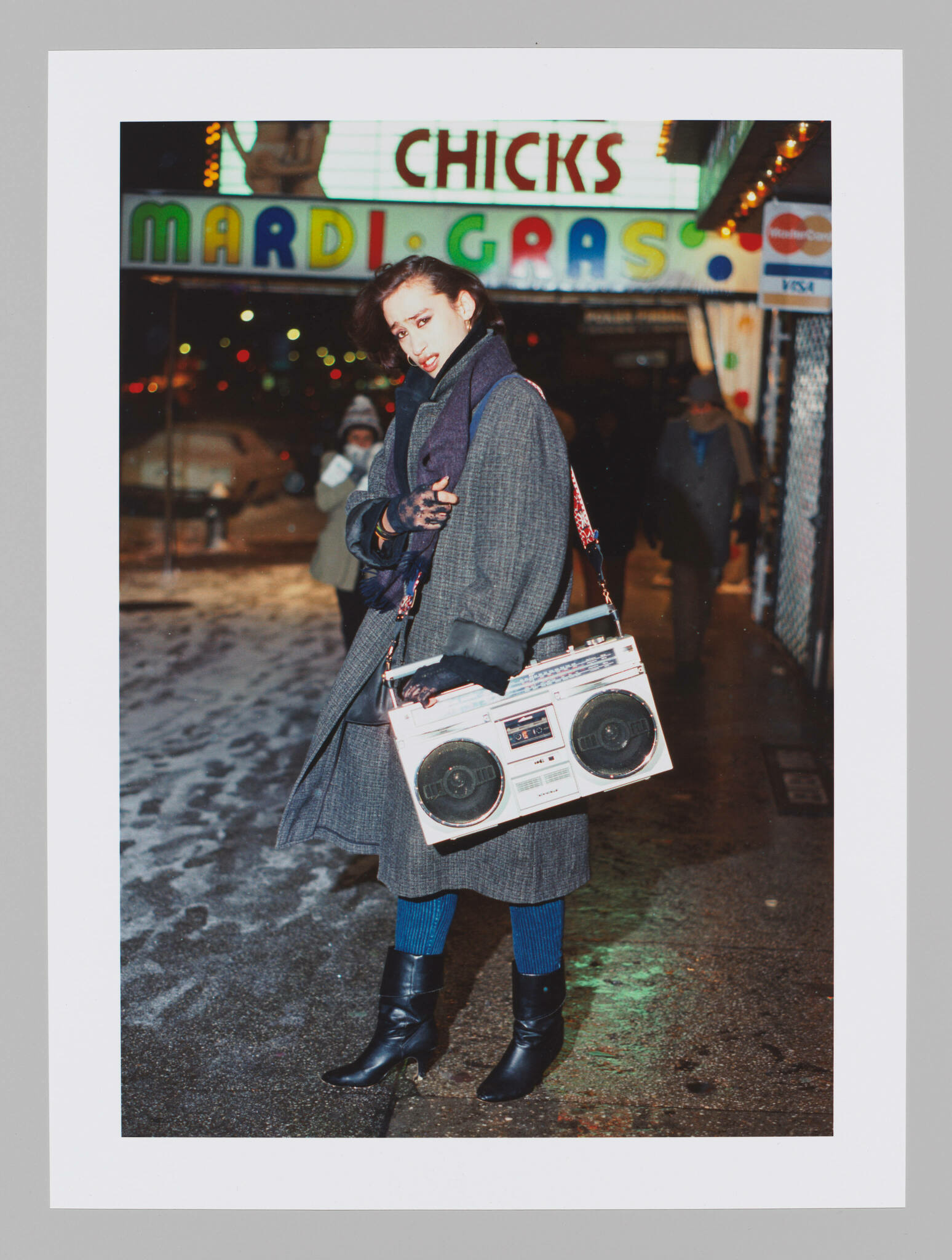 A person stands on a snowy street at night, holding a vintage boombox on their shoulder. They are wearing a long gray coat, black boots, and blue tights, with a scarf wrapped around their neck. Behind them, a neon sign reads "CHICKS MARDI GRAS," indicating a festive or party location. The individual is glancing back at the camera with a playful expression.