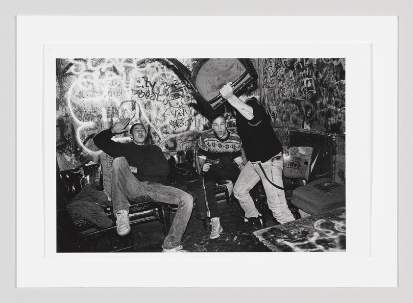 A black and white photograph capturing three young men in a graffiti-covered room. The man on the left is seated with his hand on his head, appearing surprised or shielding himself, while the man in the center is seated with a microphone, mouth open as if shouting or singing. The man on the right is standing, energetically raising a chair above his head. The chaotic environment suggests a punk or underground music scene.