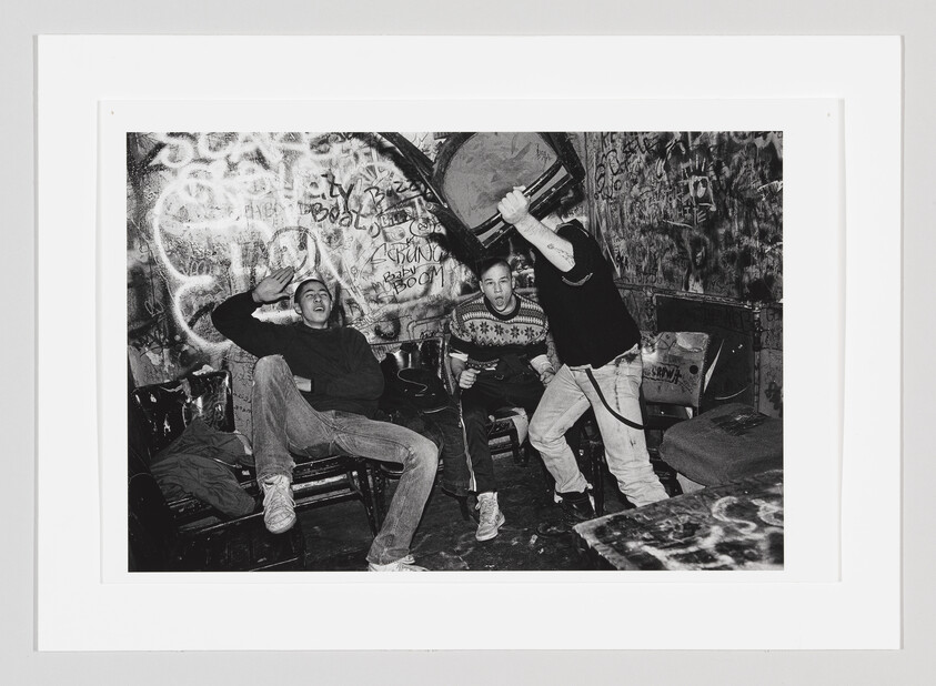 Three young men in a graffiti-covered room, one raising a chair while others react.