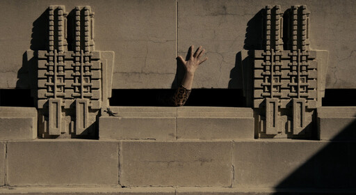A single hand in a leopard-print sleeve reaches out from a narrow gap in a concrete wall.