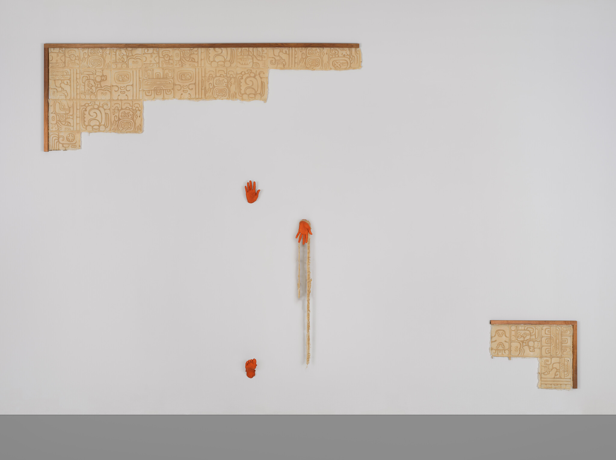 A white wall with terracotta inscribed tiles on opposite corners. In the center are two orange hands and one orange foot.