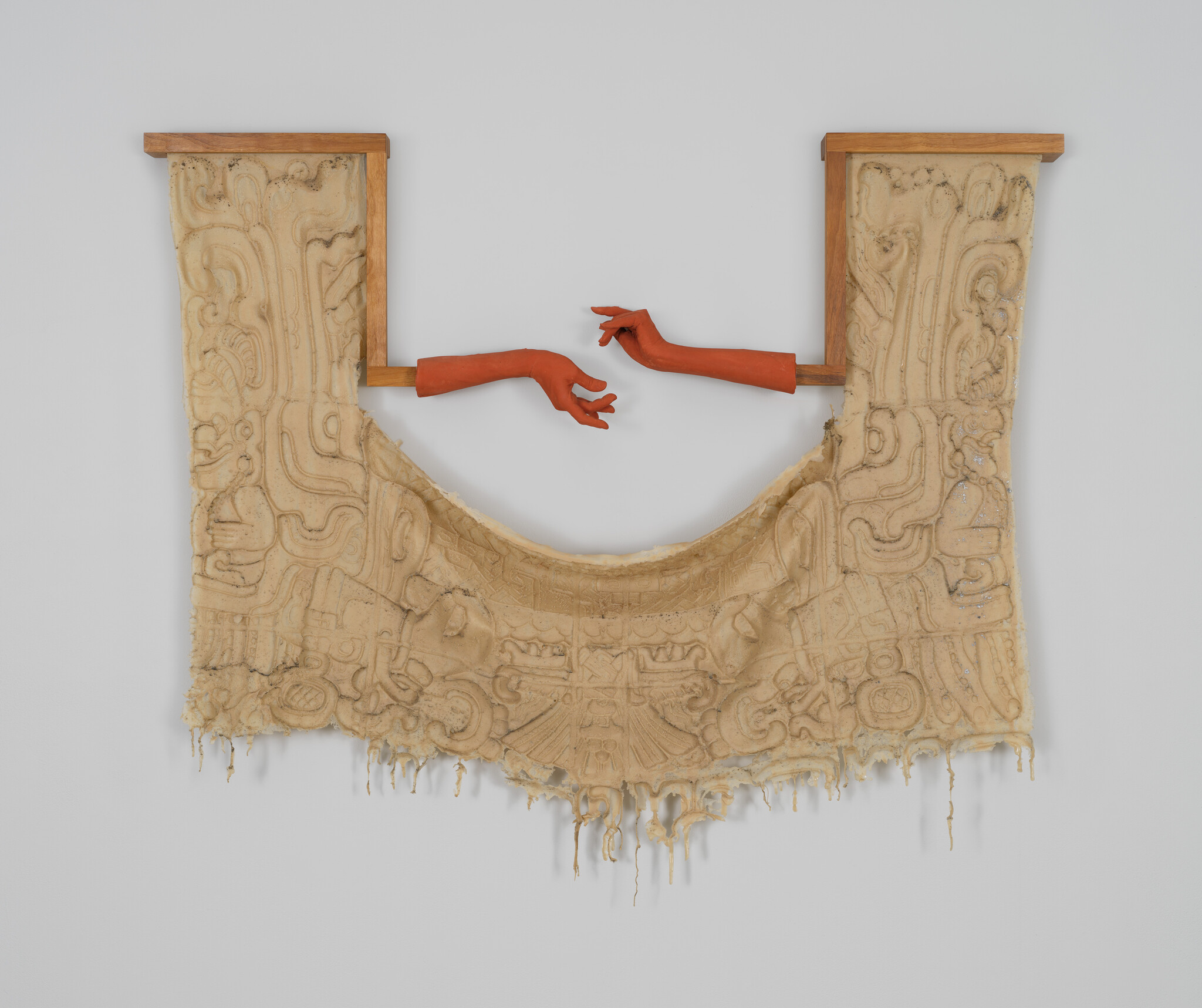 Two orange sculpted hands reach toward each other above a carved, draping beige textile wall hanging.