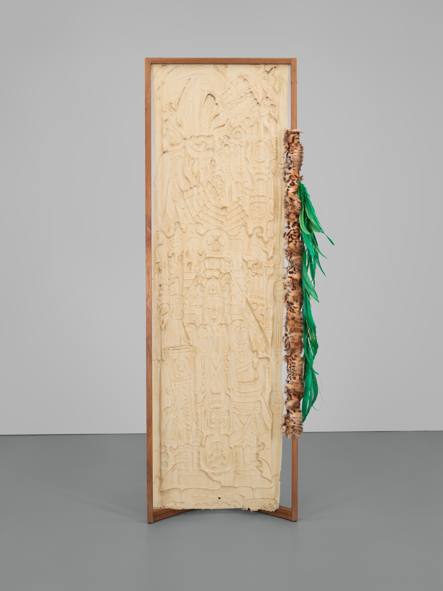 Tall carved wooden panel in a narrow frame with spotted fur trim and green feathers.