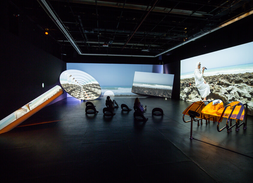 People sit on round seats in a dark room watching large screens showing beach and ocean scenes.