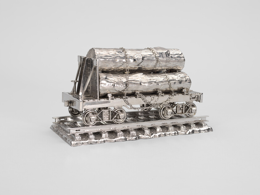 A silver model train car carries two large logs secured with chains, sitting on a short section of track.