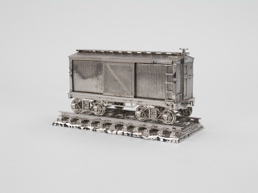 A small, shiny metal model of a boxcar train sits on a short section of metal railroad track.