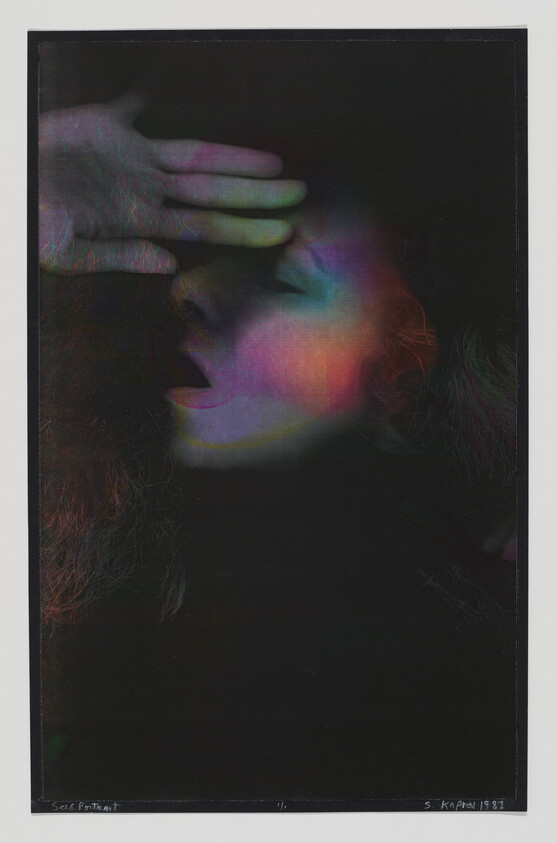 Woman in profile shields her eyes with a hand while colorful light falls across her face.